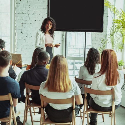 Female african-american speaker giving presentation in hall at workshop. Audience or conference hall. Rear view of participants in audience. Conference event, training. Education, diversity, inclusive concept.